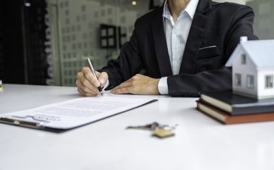 Un agent immobilier en costume signe un contrat sur un bureau avec une maquette de bâtiment et des clés.
