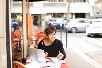 Femme assise à une table de café en extérieur, travaillant sur un ordinateur portable dans un environnement urbain.