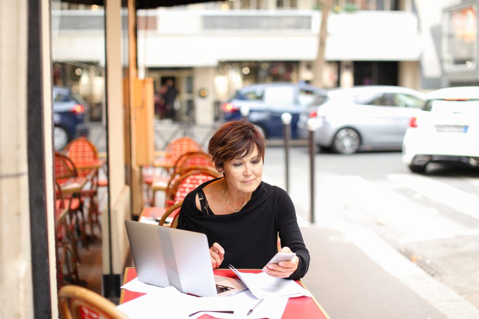 Femme assise à une table de café en extérieur, travaillant sur un ordinateur portable dans un environnement urbain.
