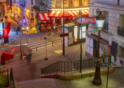 Entrée d'une station de métro éclairée dans le quartier animé de Montmartre à Paris, avec terrasses de cafés et passants.